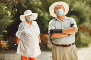 foto com arvores ao fundo e, na frente, há dois senhores, uma senhora no canto esquerdo usando roupas claras, chapéu e máscara, no outro lado há um senhor com camiseta azul clara, cinto, berumada, usando também um chapéu e máscara, ele está de braços cruzados, ilustrando idoso com covid
